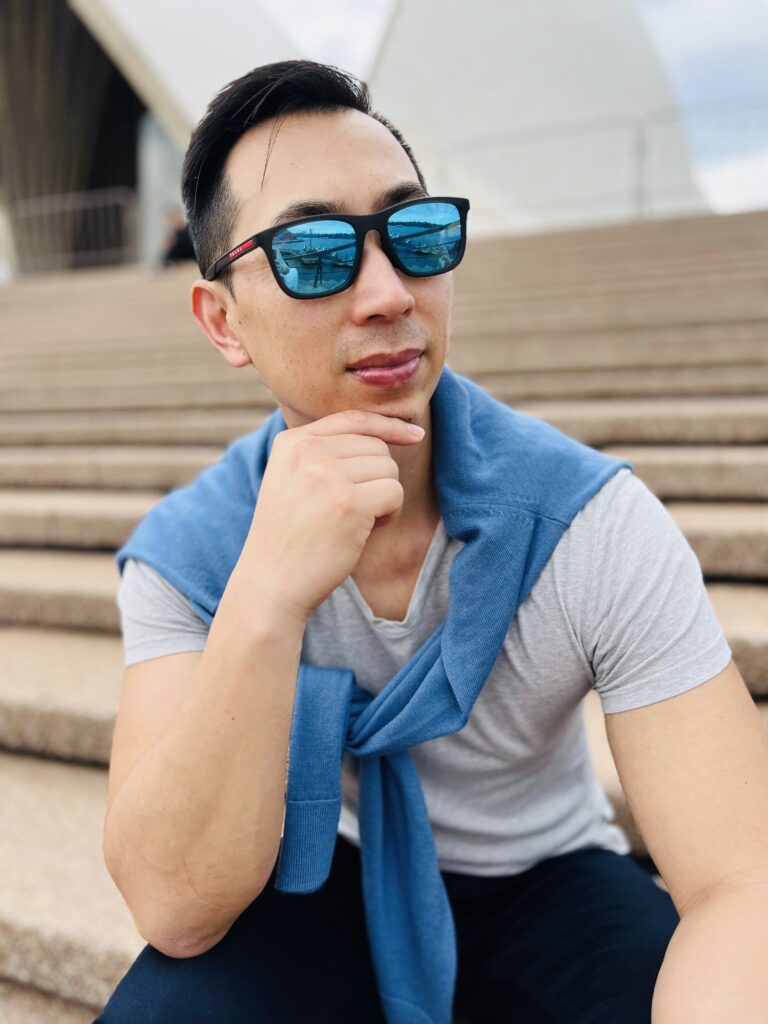 Male sitting on the steps of the Opera House Sydney wearing sunnies and a blue sweater over his shoulders